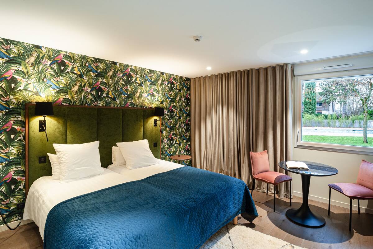 Quarto de hotel com uma cama grande, uma mesa pequena com cadeiras, uma janela com cortinas e um papel de parede tropical.