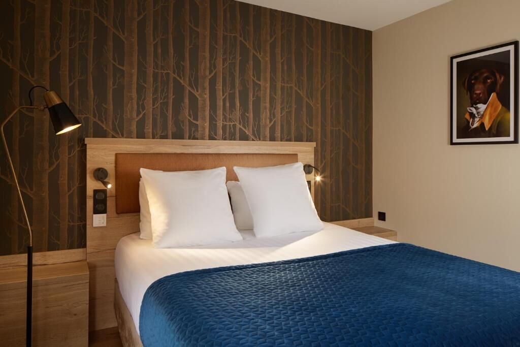 Ein Hotelzimmer mit einem hölzernen Kopfteil, einer blauen gesteppten Bettdecke, einer Wandleuchte und einem gerahmten Bild an der Wand.