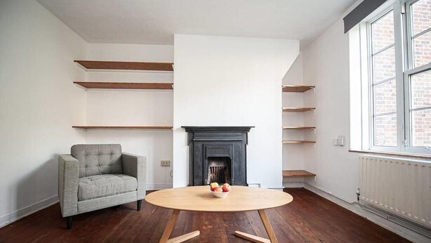 A room with a fireplace, wooden shelves, a grey armchair, and a round wooden table with a bowl of fruit. - Central London 2BR Apartment in Waterloo (London)