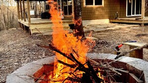 Un foyer en pierre avec un feu allumé, situé devant une cabane en bois surmontée d’un toit en métal.