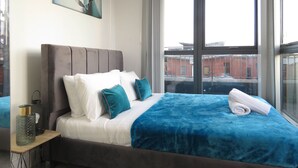 A modern bedroom with a large window, a bed with blue and white bedding, and a decorative pillow. - Tudors eSuites City Centre Apartment (Birmingham)