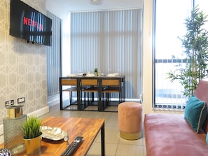 A modern living room with a flat-screen TV displaying 'NETFLIX', a dining area with a table and chairs, a coffee table with a remote control, and a large window with blinds. - Tudors eSuites City Centre Apartment (Birmingham)