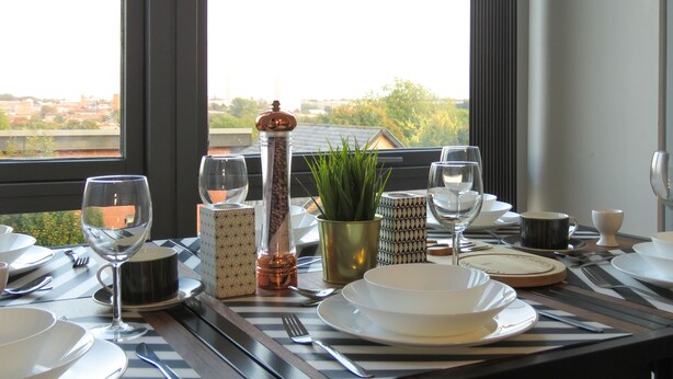 A dining table set with plates, glasses, and cutlery, with a view of a cityscape through large windows. - Tudors eSuites City Centre Apartment (Birmingham)