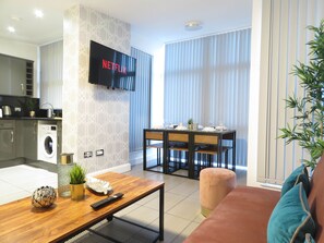 A modern living room with a flat-screen TV mounted on the wall, a dining area with a table set for four, and a kitchen area with a washing machine. - Tudors eSuites City Centre Apartment (Birmingham)