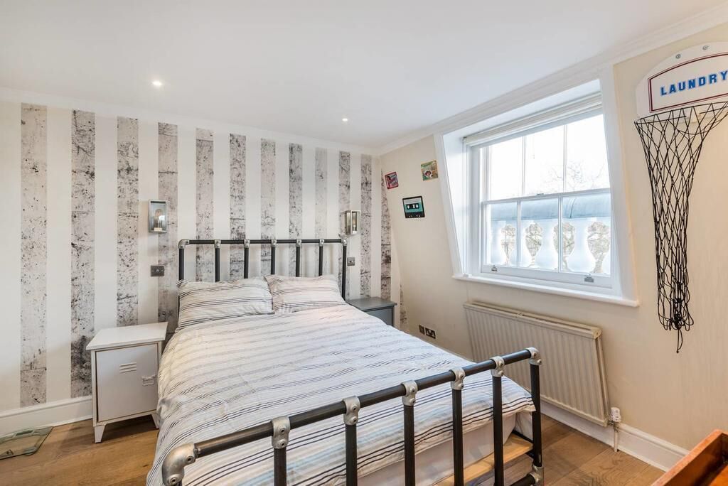 A bedroom with a bed, a nightstand, a radiator, a window, and a laundry basket on the wall.