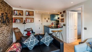 A compact living space with a black sofa, patterned cushions, a brick wall, a bookshelf, a small kitchen area, and a bicycle. - Lovely 1BR Flat Walk to Hyde Park (London)