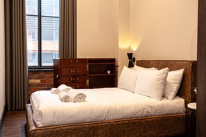 A neatly made bed with white linens and rolled towels, a wooden dresser with multiple drawers, a small bedside table with a lamp, and a brick wall visible through a window with curtains. - Portland Street Apartments (Manchester)