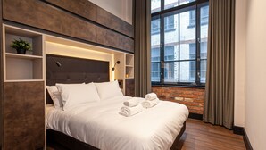 Quarto de hotel moderno com uma cama grande, estantes embutidas, um vaso de planta e vista da cidade através de uma grande janela.