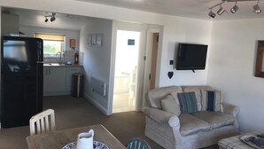 Un espacio habitable compacto con kitchenette, una zona de comedor con mesa y sillas, un sofá y un televisor montado en la pared.