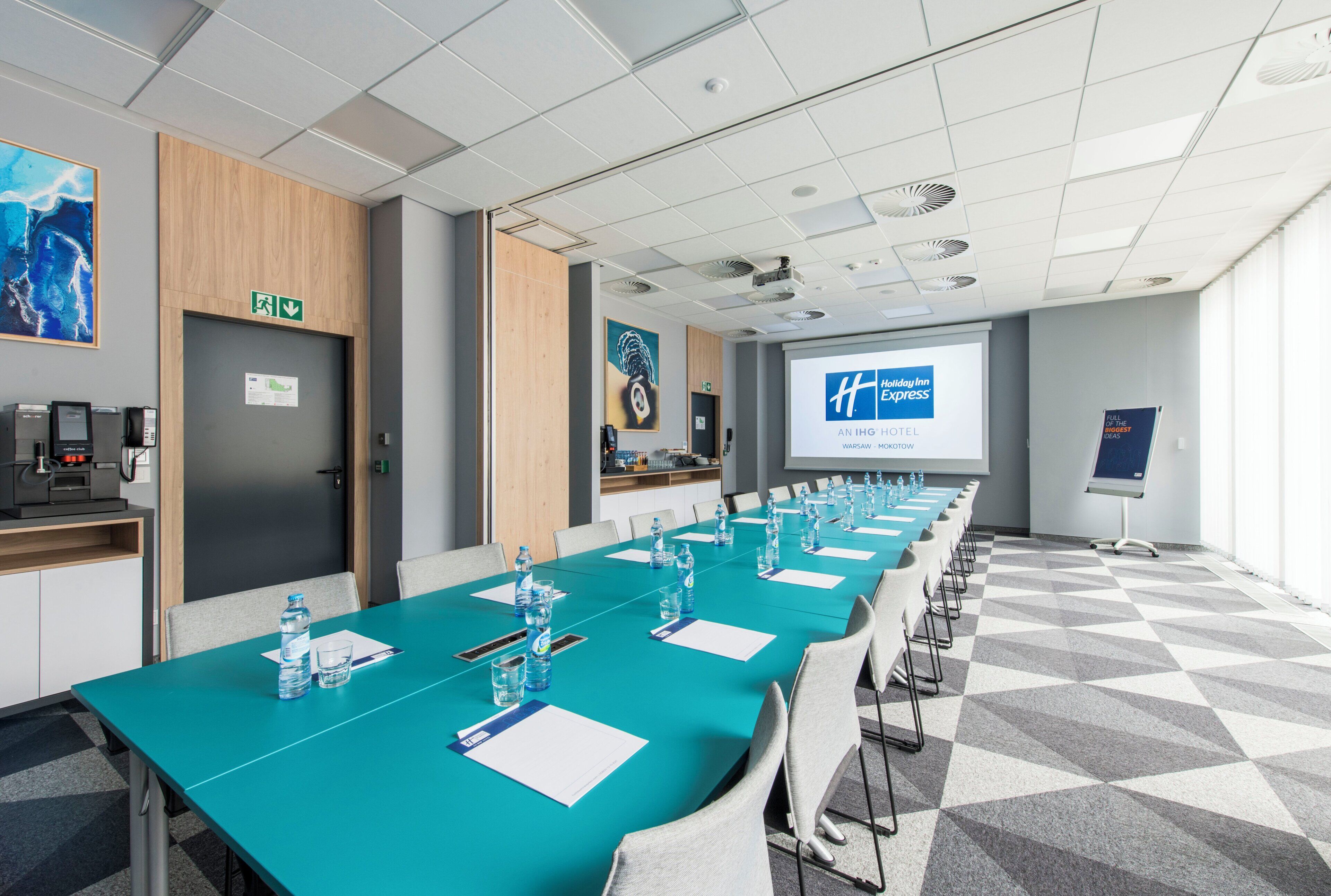A conference room with a long table, chairs, a coffee machine, and a projector screen displaying a hotel name.