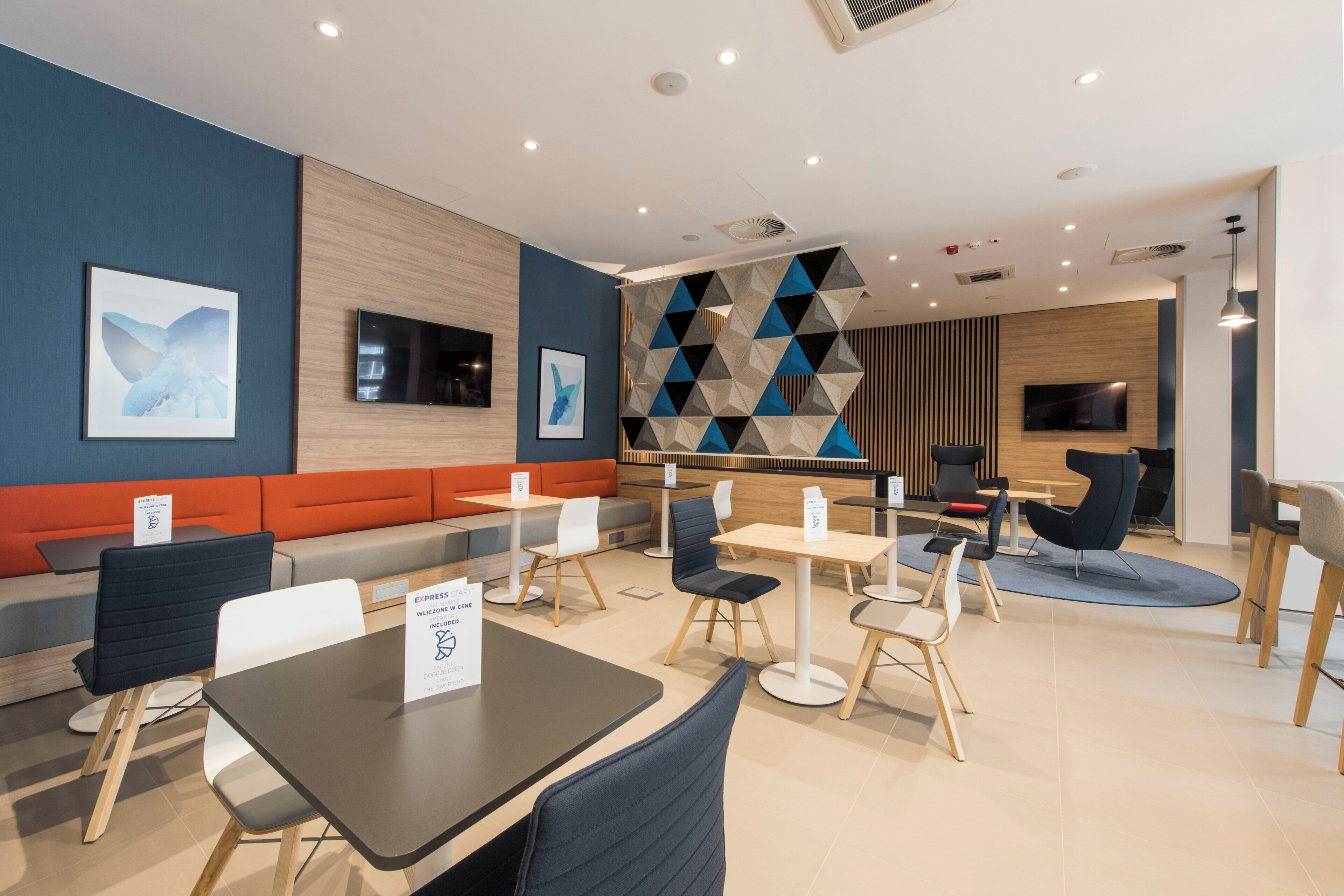 A modern cafe with seating arrangements including tables and chairs, a wall-mounted TV, and a geometric patterned wall.