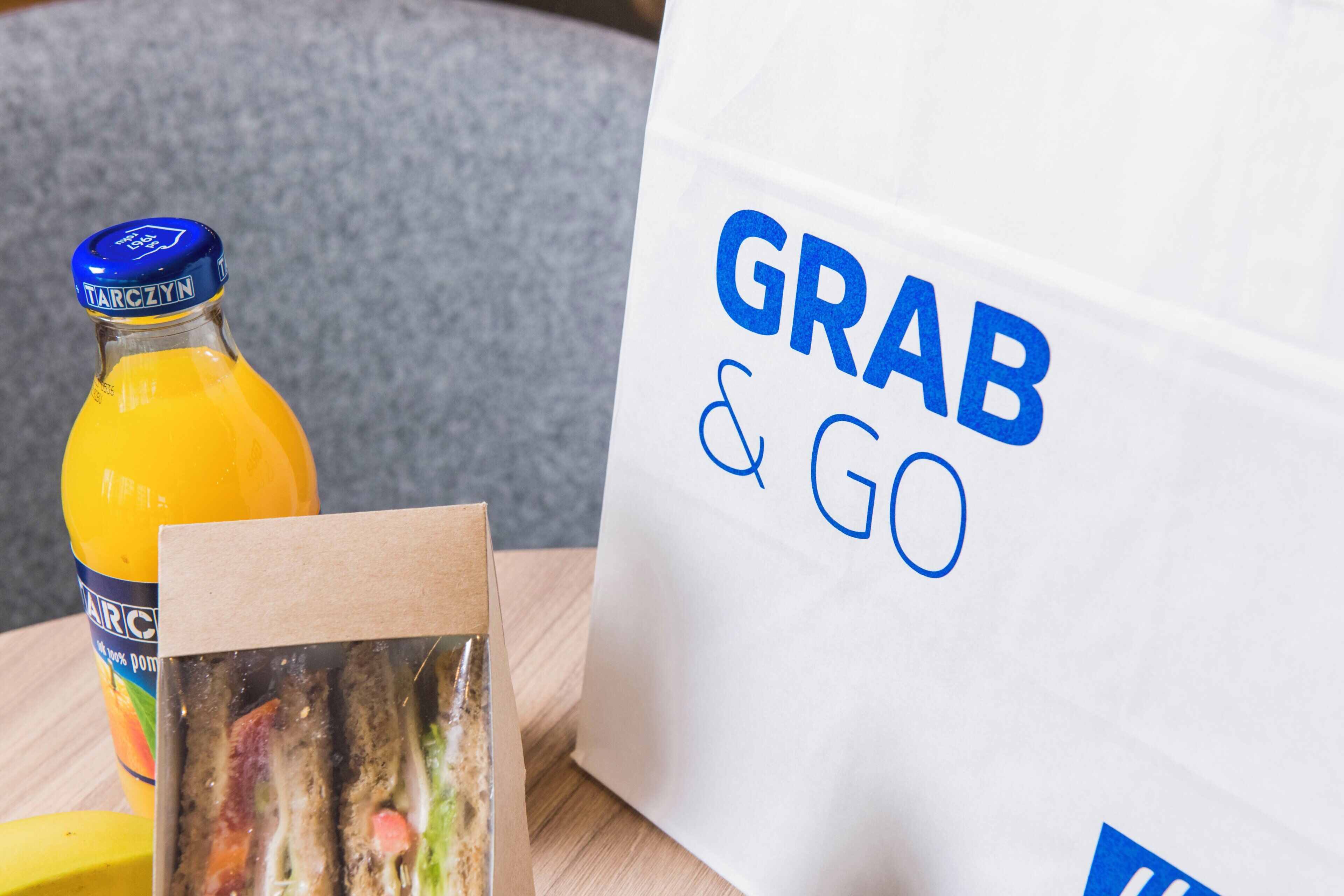 Une bouteille de jus d’orange, un sandwich et un sac en papier blanc sur lequel est imprimé le texte « À EMPORTER ».