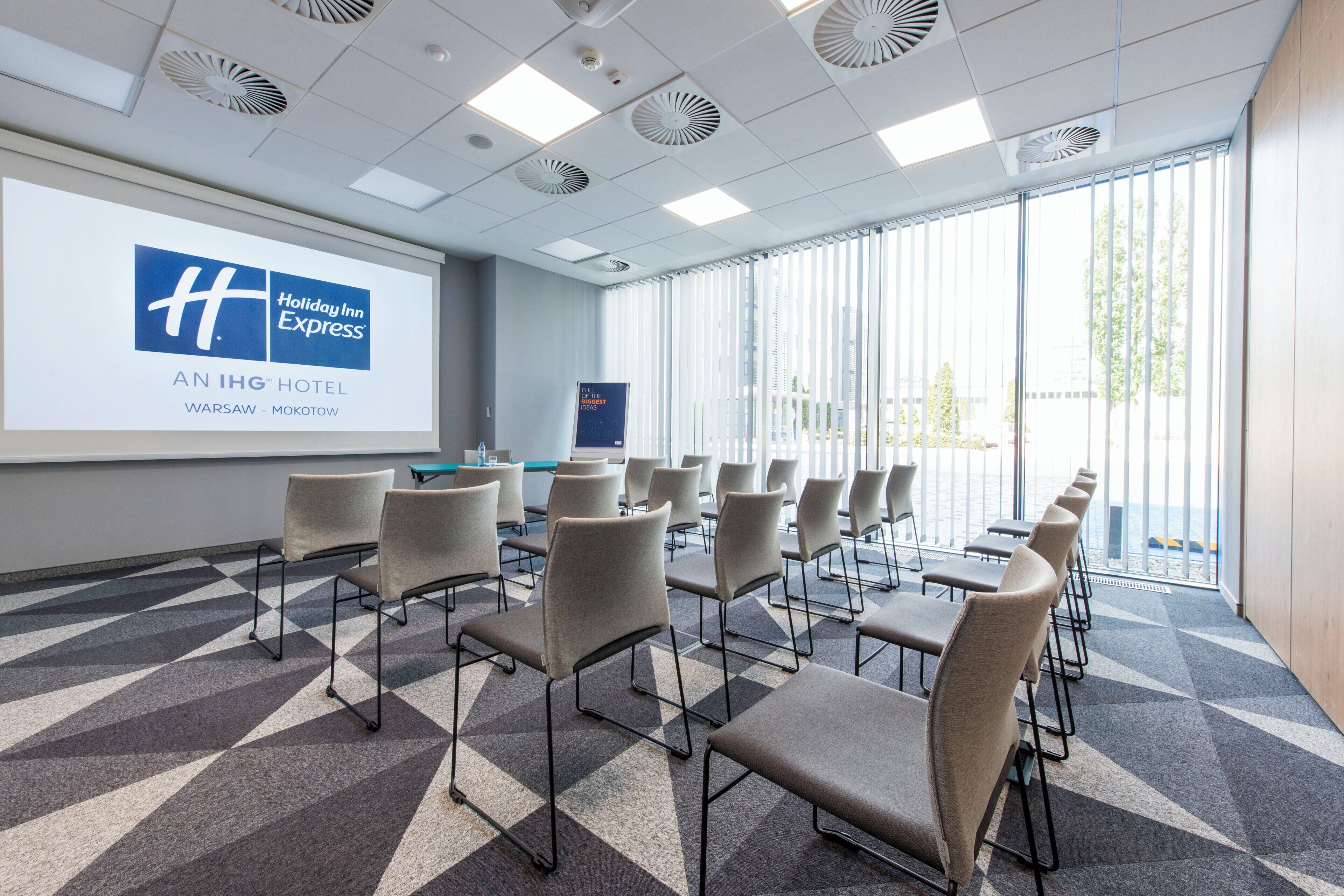 A conference room with a projection screen displaying a hotel logo, arranged seating, and large windows.