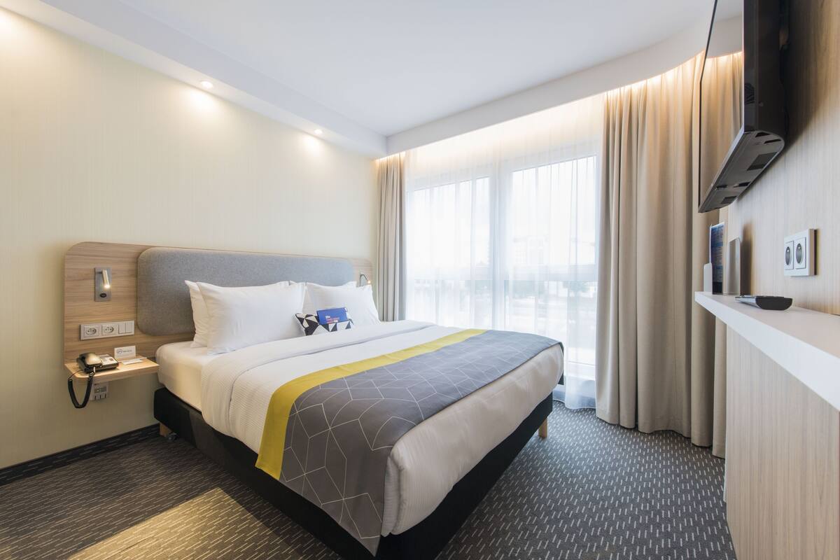 A hotel room with a bed, a bedside table with a phone, a flat-screen TV mounted on the wall, and a large window with curtains.