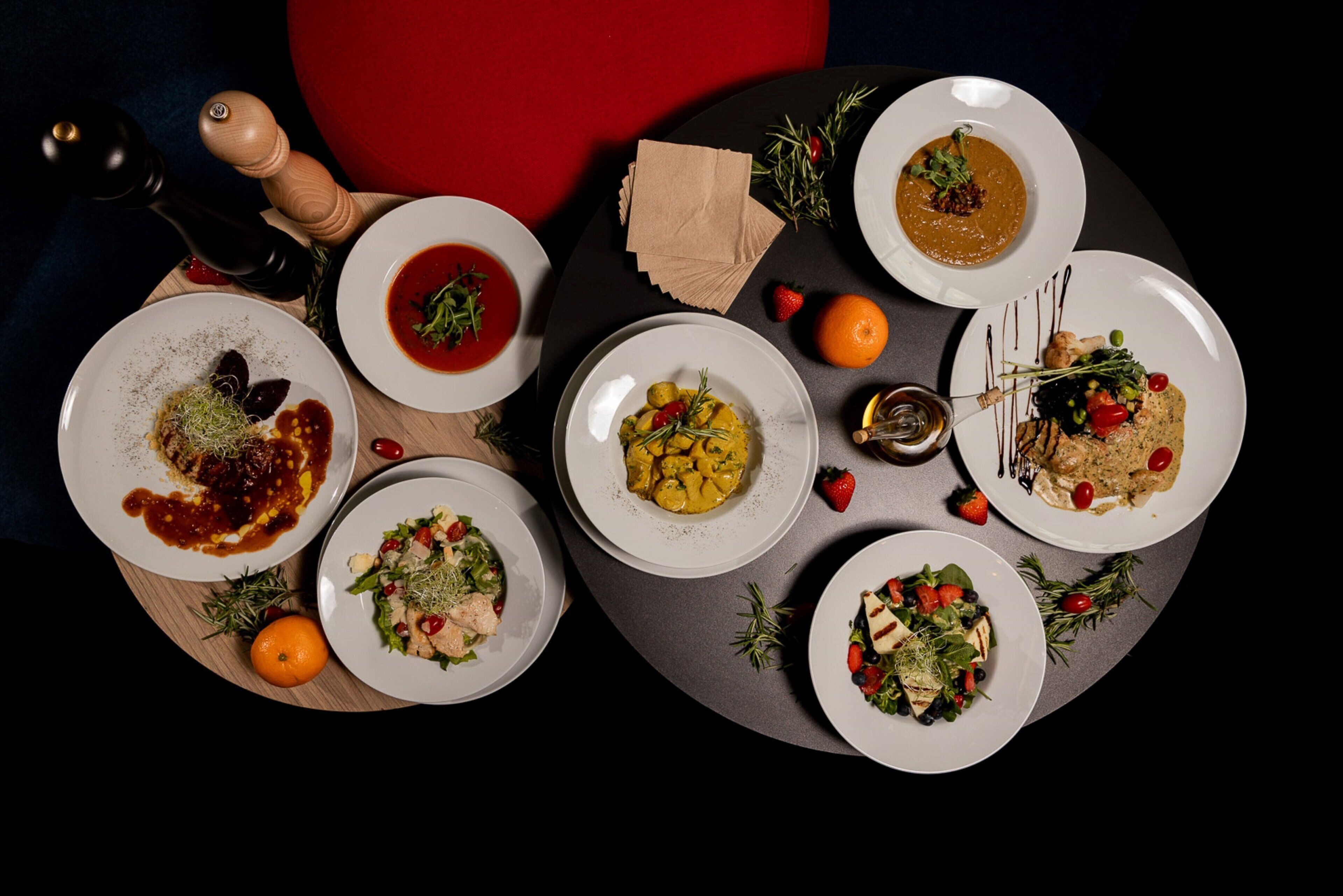 A table with various dishes, including a bowl of soup, a plate with a salad, and a plate with a dessert. There are also two oranges and a pepper grinder on the table.
