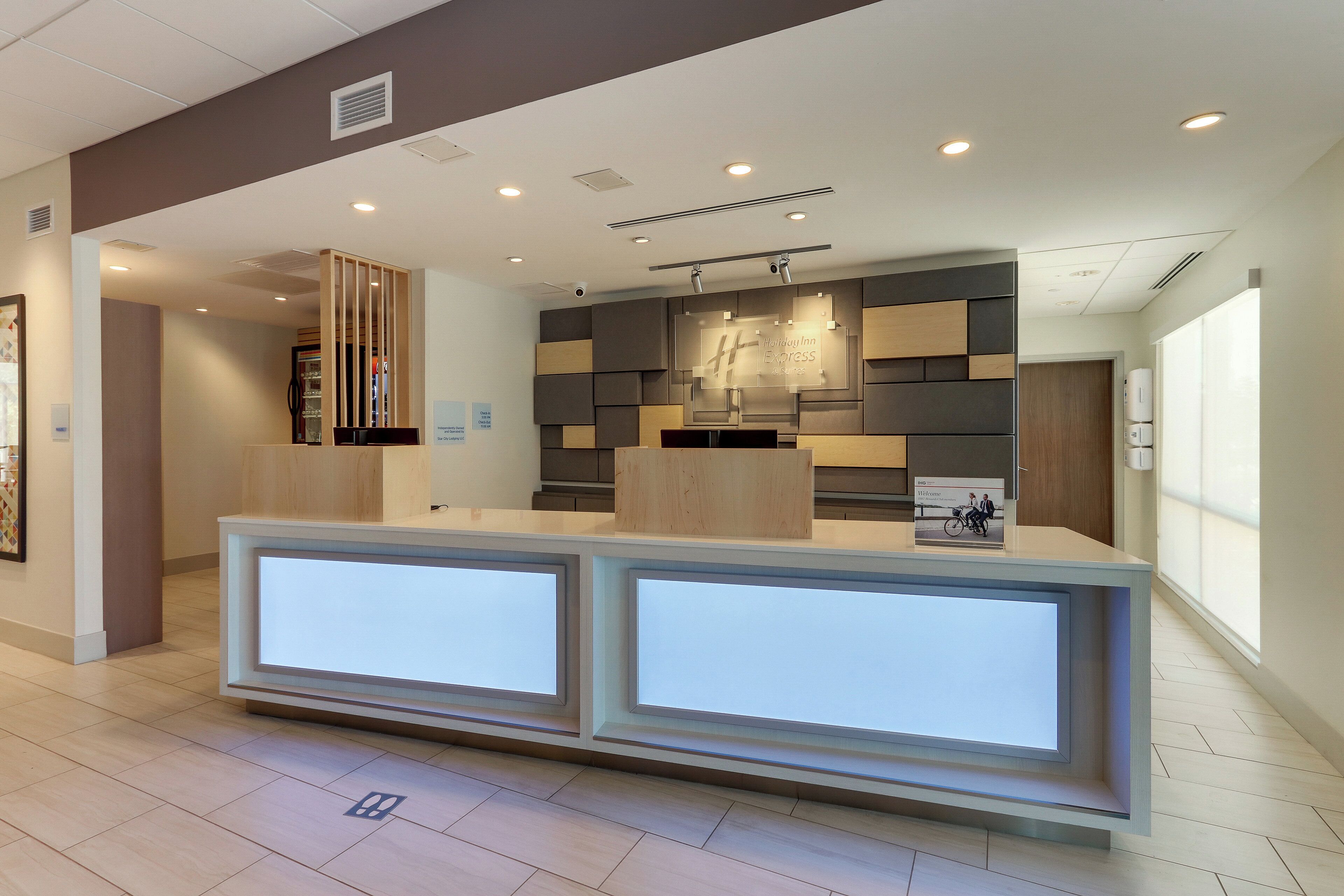 A hotel reception area with a modern design, featuring a reception desk, a wall-mounted information board, and a visible ceiling with recessed lighting.