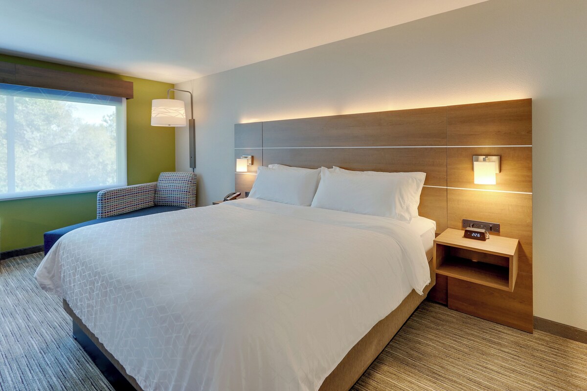 A hotel room with a large bed, a nightstand with a clock, a wall-mounted lamp, and a view of trees through a window.