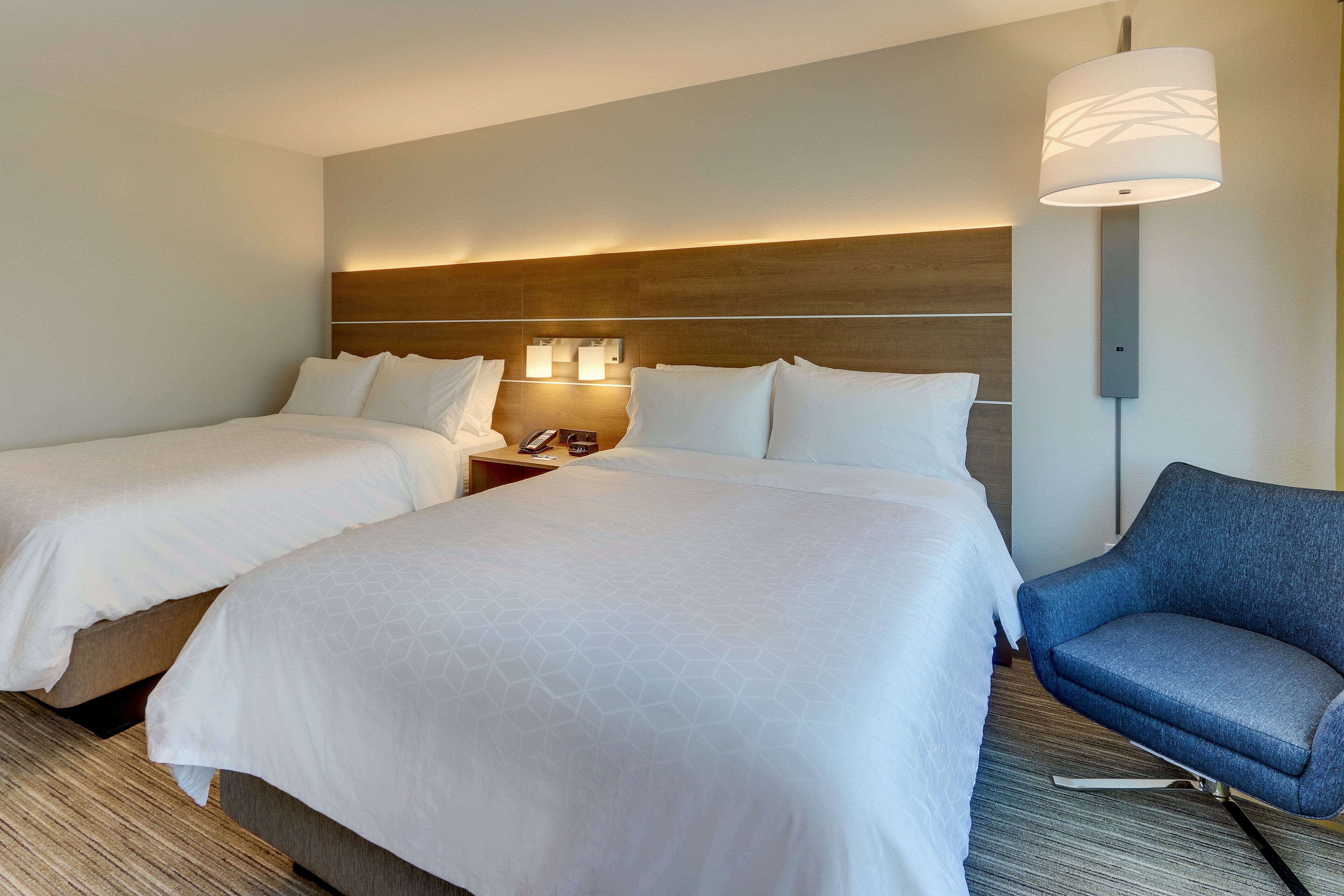 A hotel room with two beds, a nightstand, a lamp, and a blue chair.
