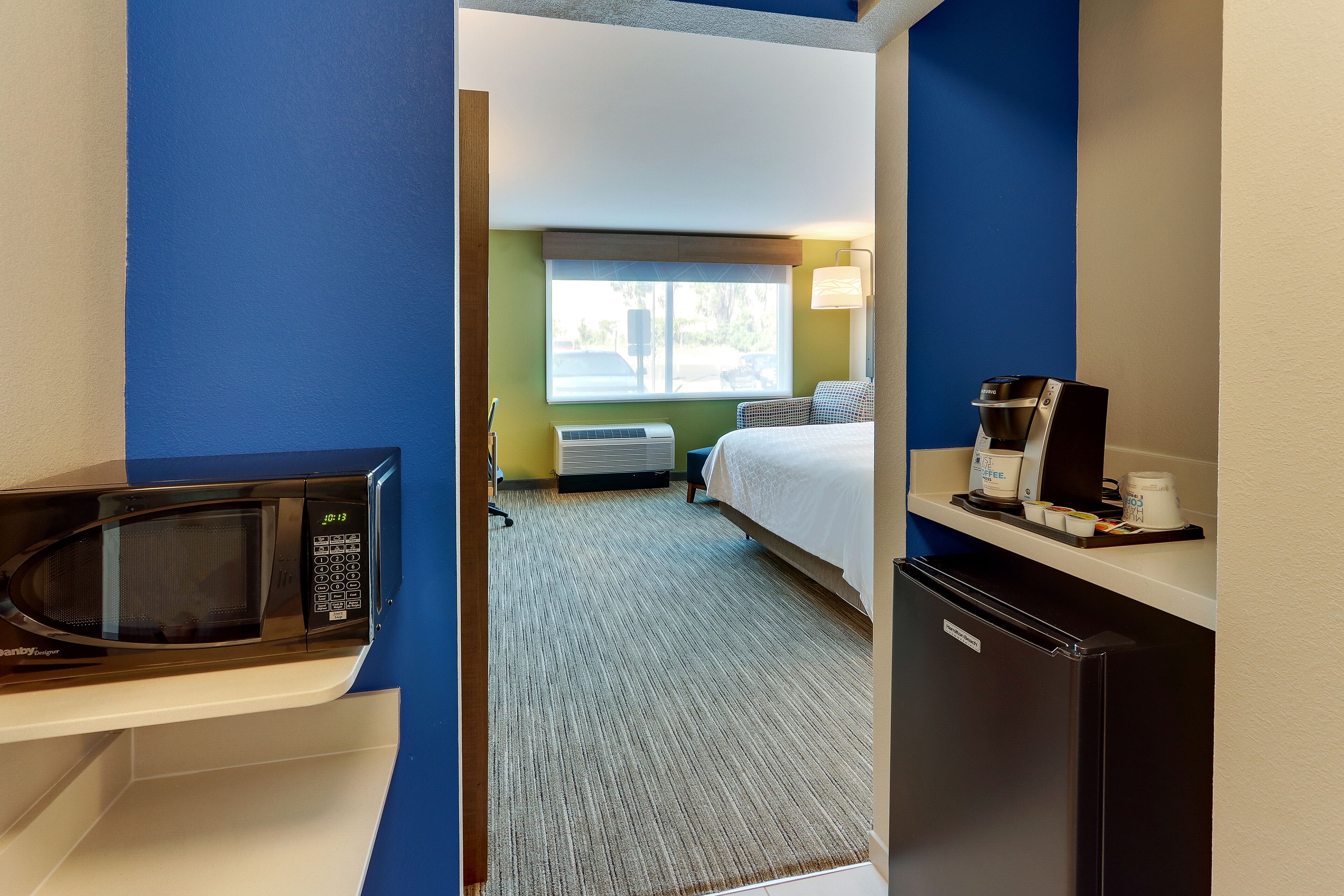 A hotel room with a microwave, a refrigerator, a bed, a window, and a coffee maker on the counter.