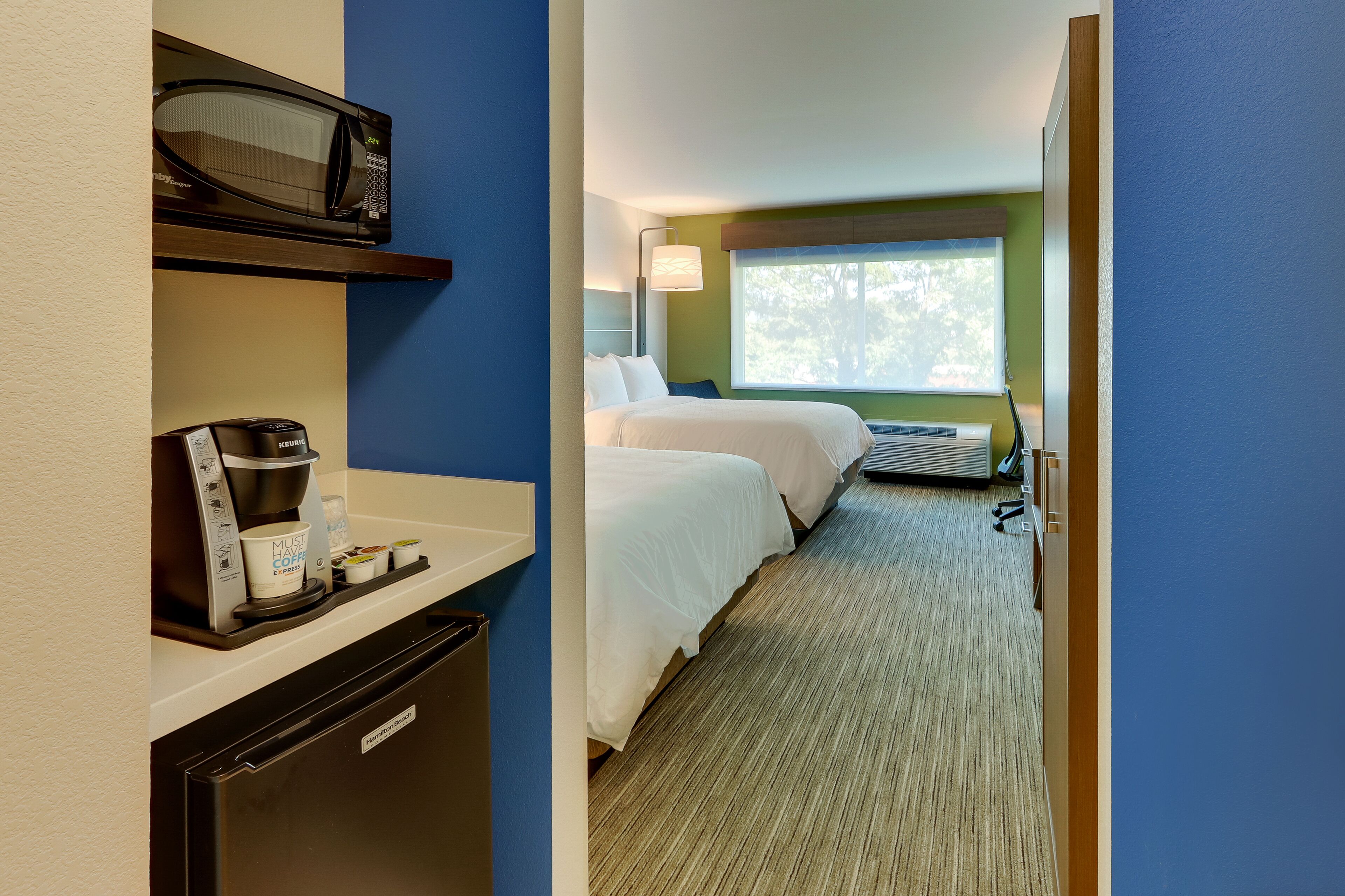 A hotel room with a kitchenette, a microwave, a coffee maker, a bed, a window with a view, and a bedside table.