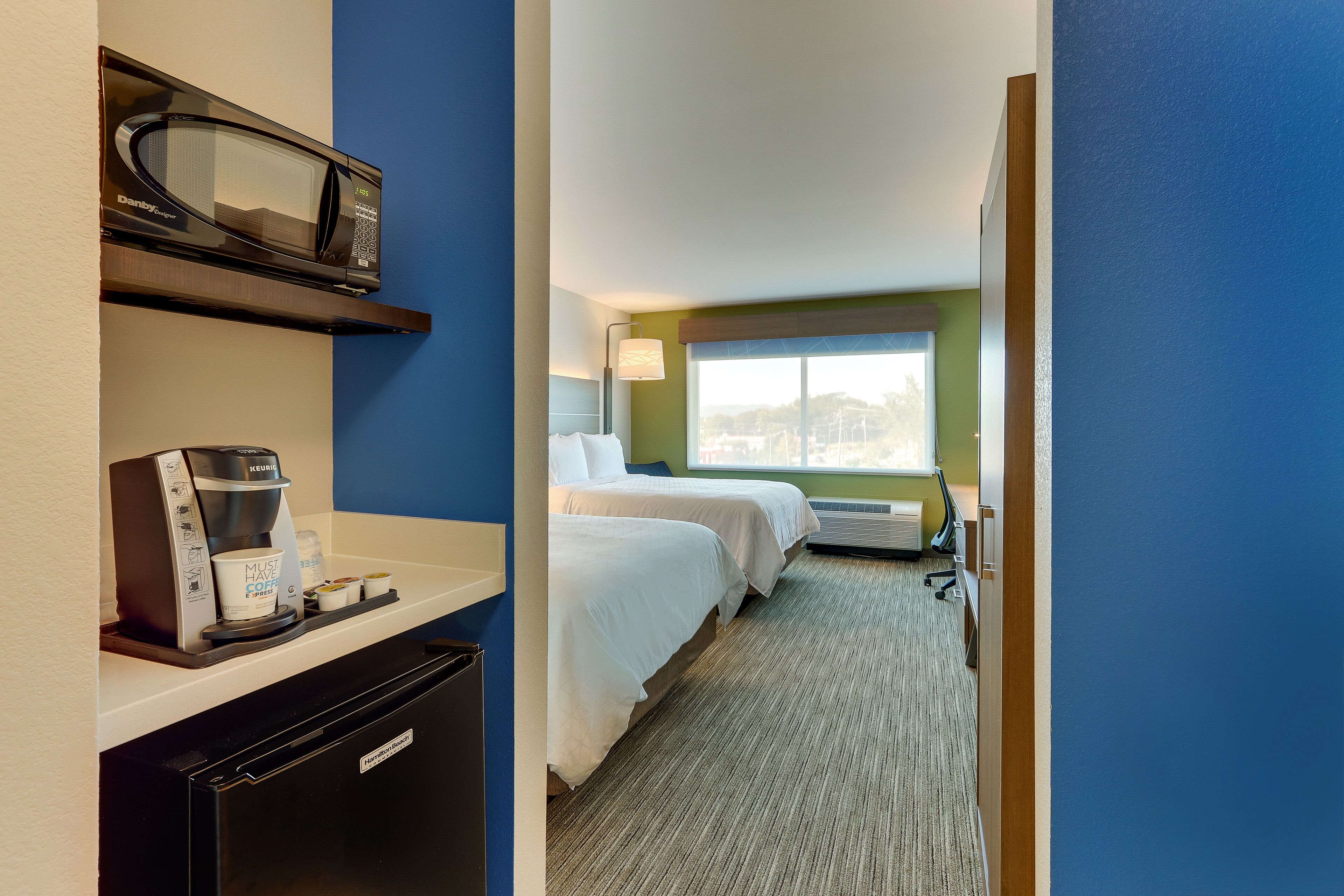 A hotel room with a kitchenette, a microwave, a coffee maker, two beds, a window with a view, and a door leading to another room.