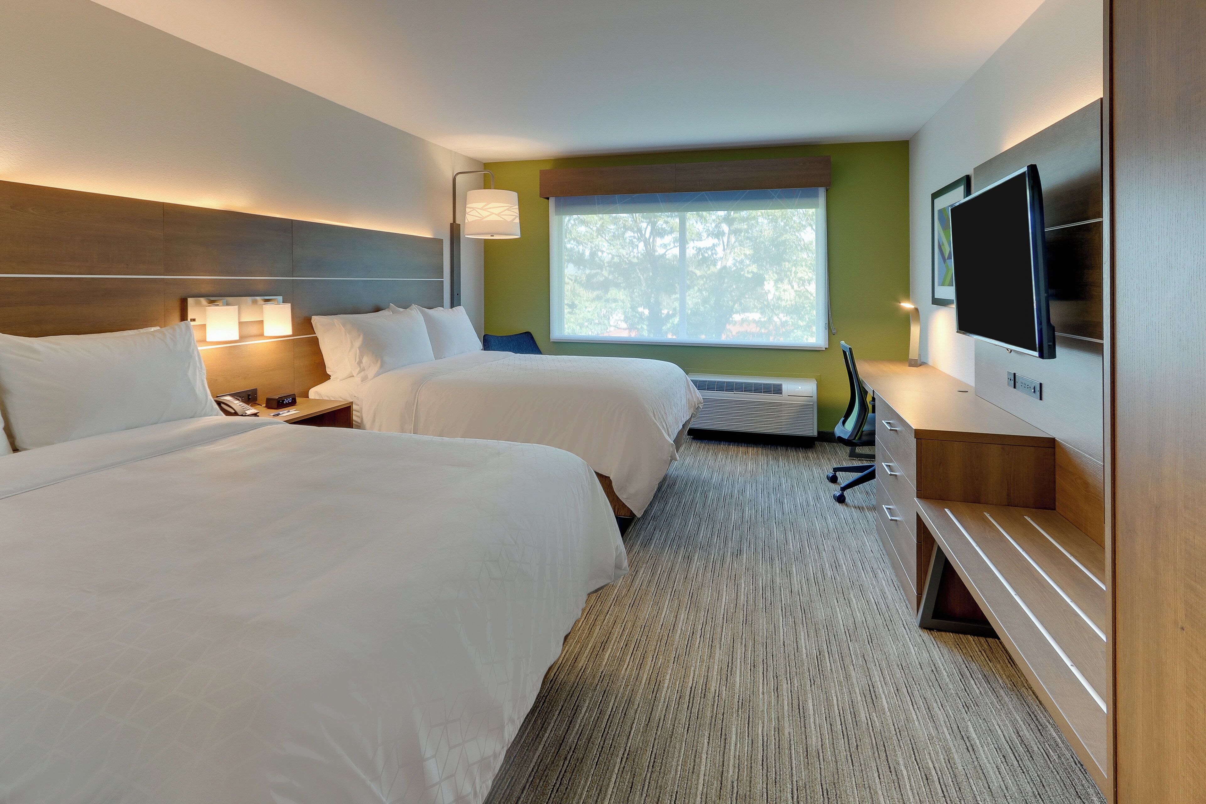 A hotel room with two beds, a TV, a desk, and a window with a view of trees.