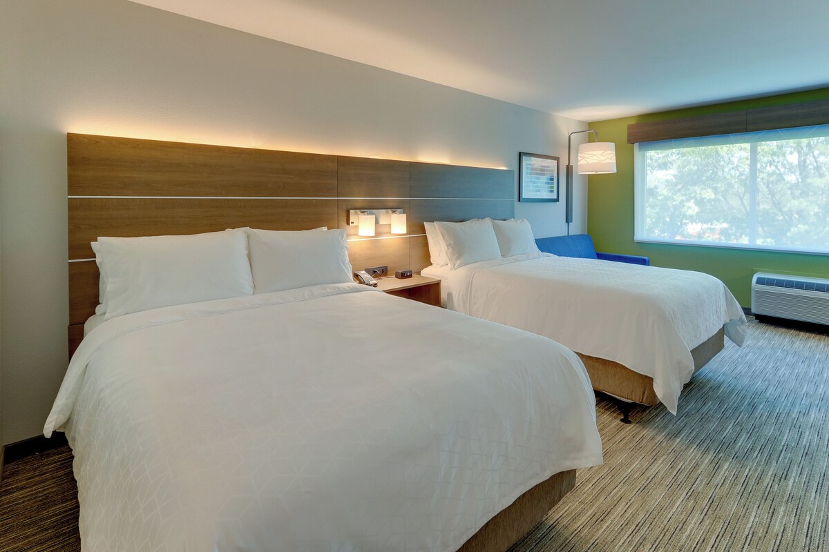 A hotel room with two beds, a nightstand, a lamp, and a window with a view.