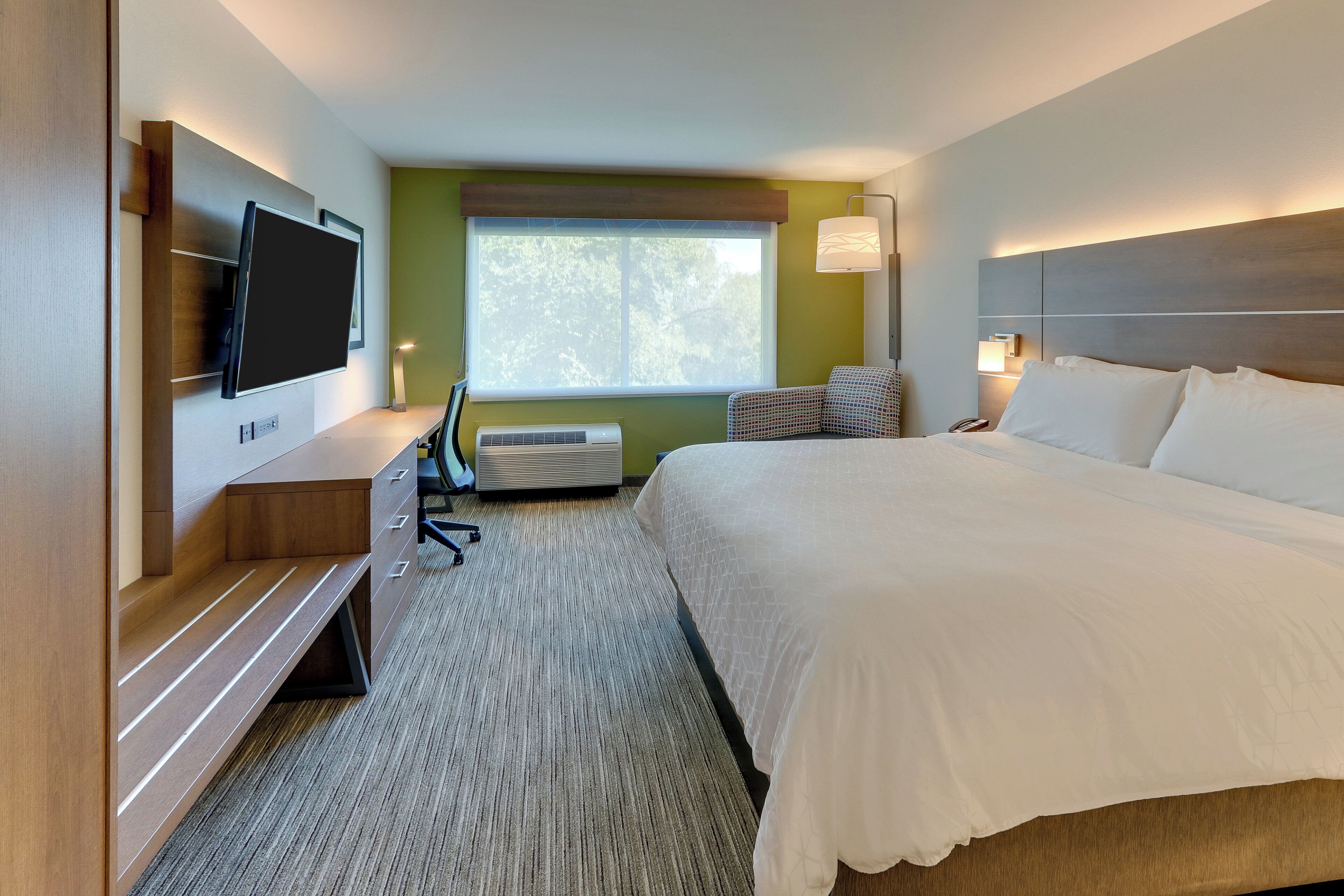 A hotel room with a bed, a desk with a chair, a flat-screen TV, a window, and a wall-mounted lamp.