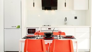 A modern kitchen with white cabinetry, a black range hood, and a dining area with a white table and orange chairs. - Paradiso III (Malevizi)