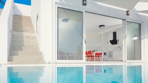 A modern pool area with clear glass walls, a kitchen visible through the glass, and a staircase leading to an upper level. - Paradiso III (Malevizi)