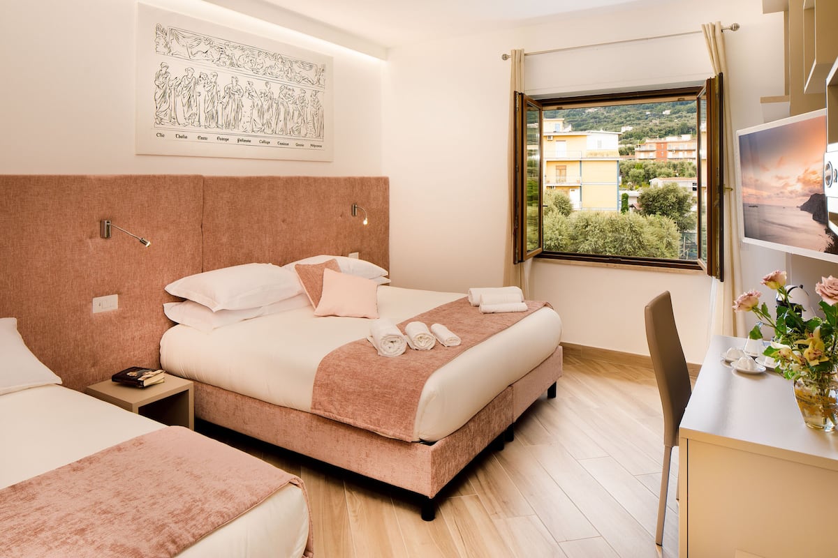 A hotel room with a large bed, a bedside table with a book, a desk with a vase of flowers, and a window with a view of buildings.