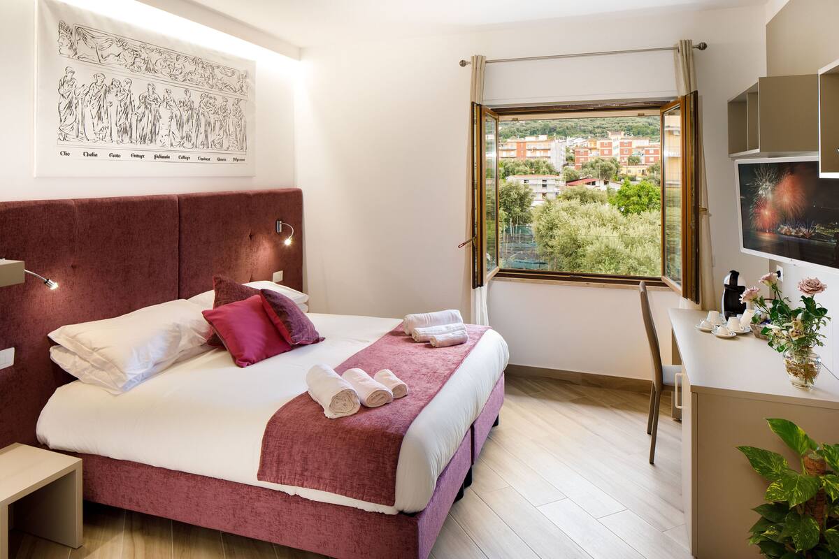 A hotel room with a large bed, a desk with a computer, a window with a view of buildings, and a framed artwork on the wall.
