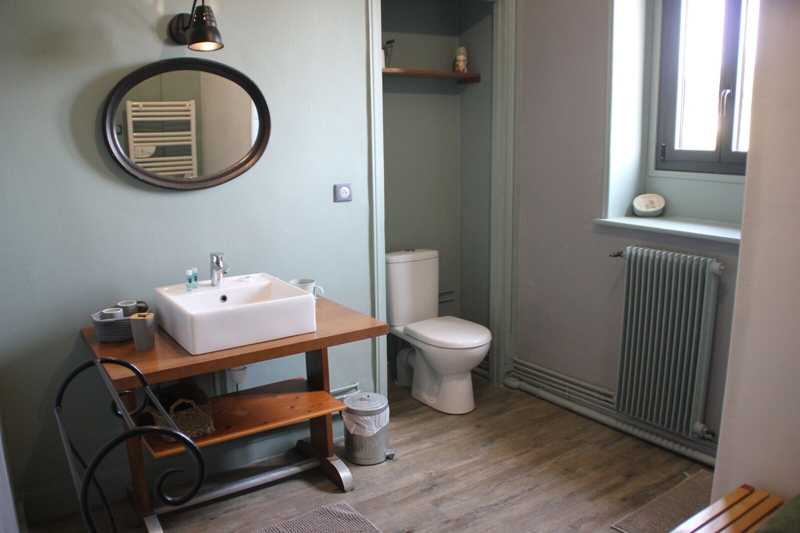 Une salle de bain avec un lavabo, des toilettes et un radiateur, tous de couleur blanche. Un miroir est accroché au mur et une fenêtre se trouve au-dessus des toilettes.