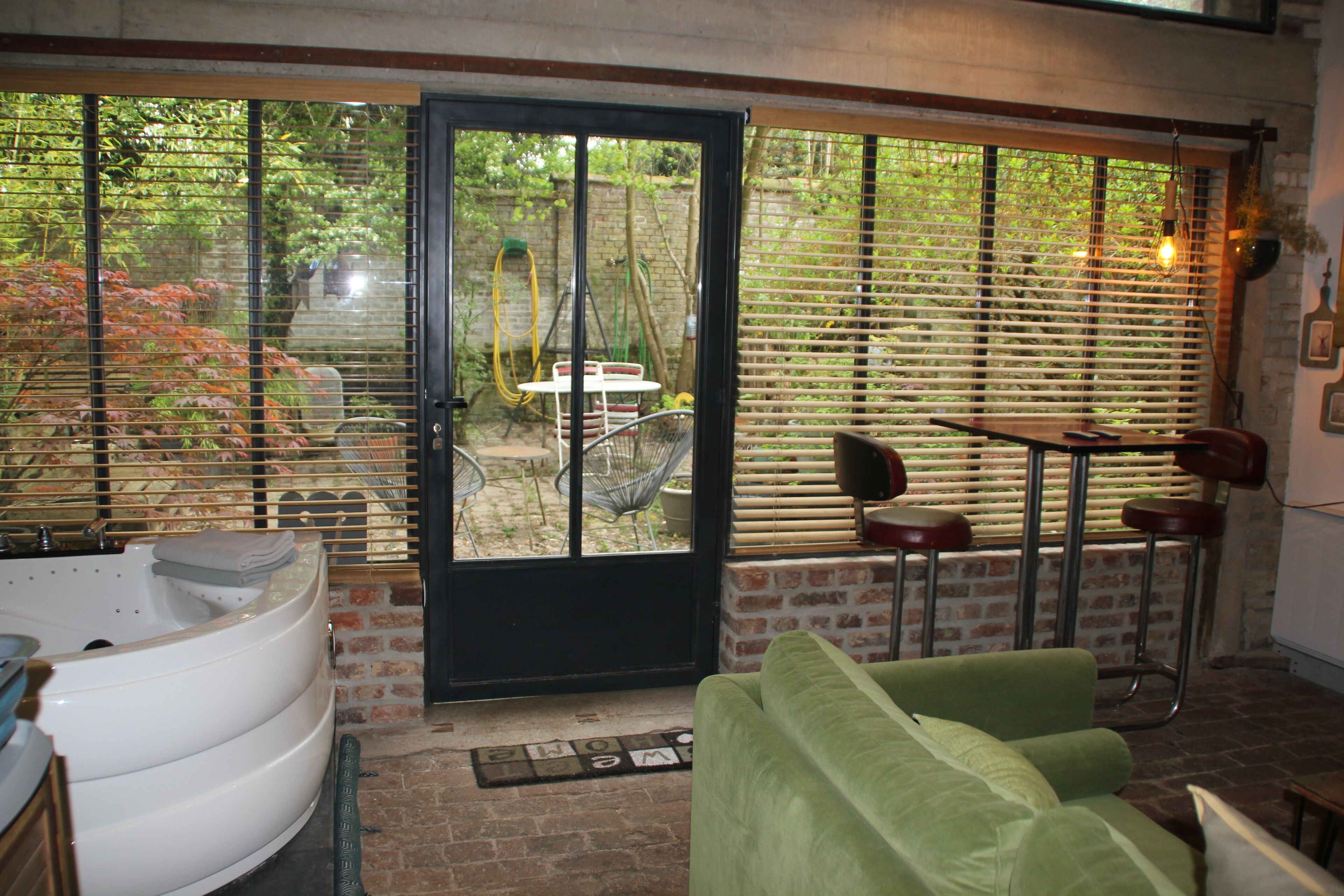 A room with a spa tub, a green sofa, a dining table with chairs, and a view through a door with blinds.