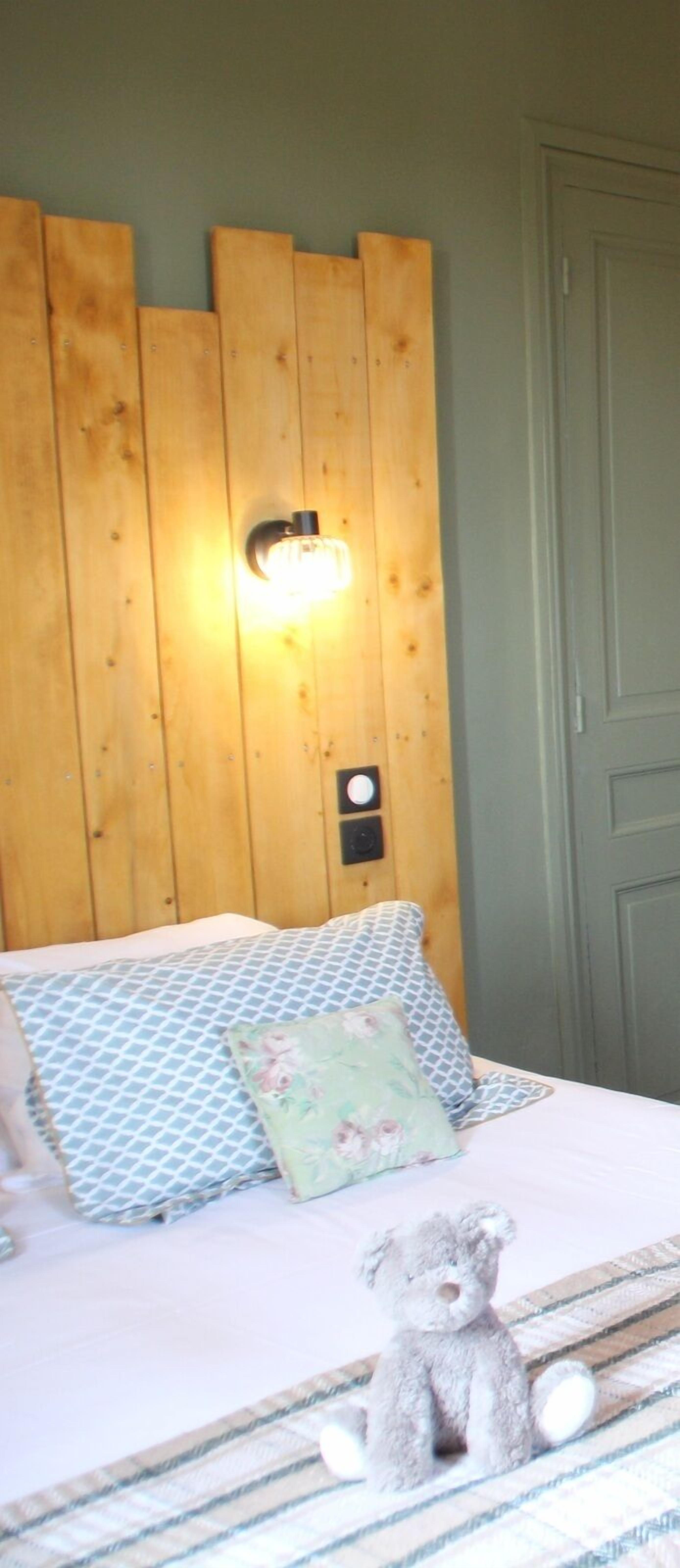 Un dormitorio con cabecera de madera, cama con ropa de cama estampada, un osito de peluche en la cama y una lámpara de pared.