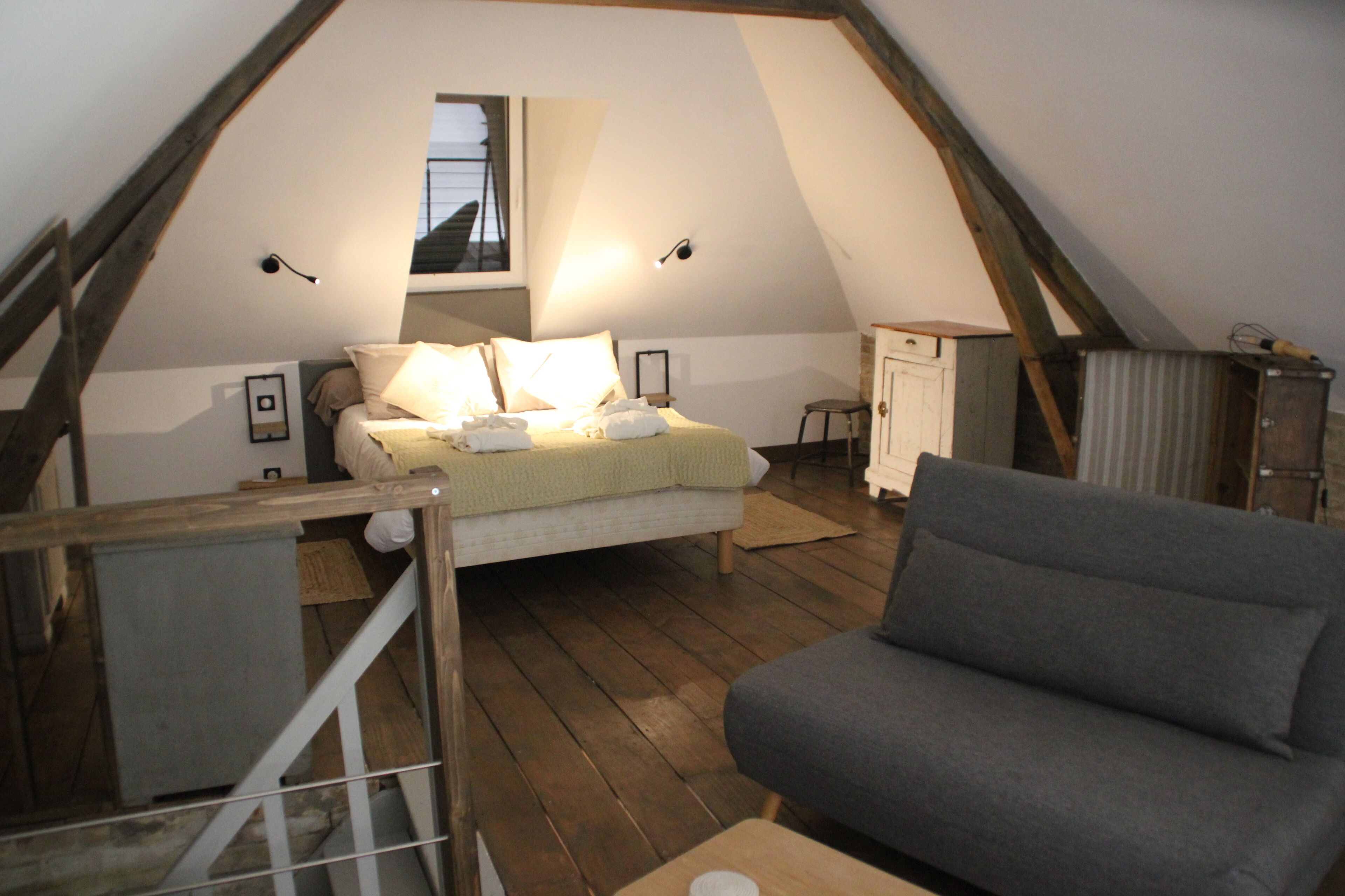 A cozy attic bedroom with a bed, a gray sofa, wooden flooring, and a small window.