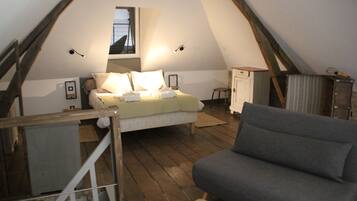 A cozy attic bedroom with a bed, a gray sofa, wooden flooring, and a small window.
