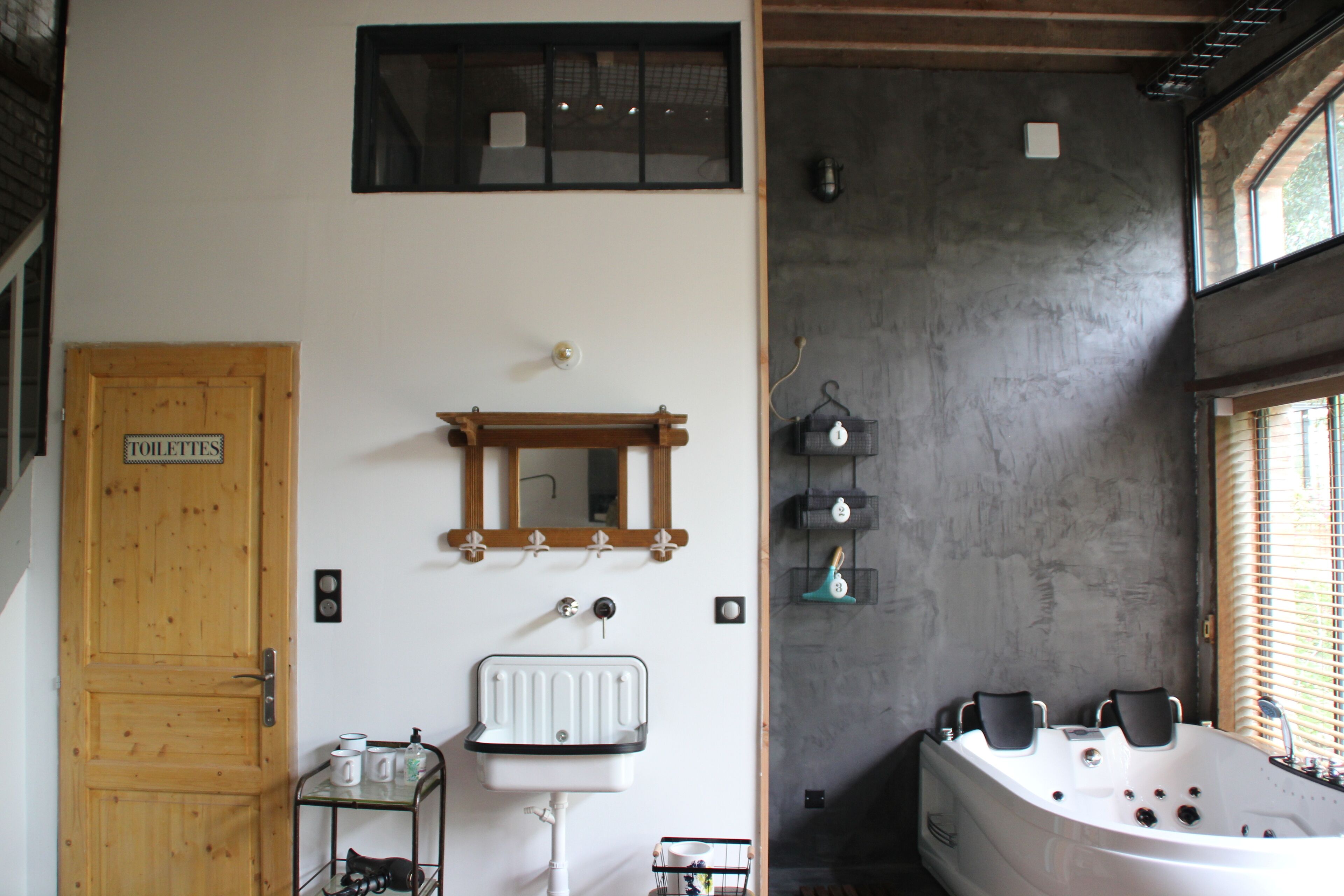 Un baño con puerta de madera que indica "TOILETTE", un lavamanos blanco, un espejo, un perchero, una zona de ducha y una bañera.