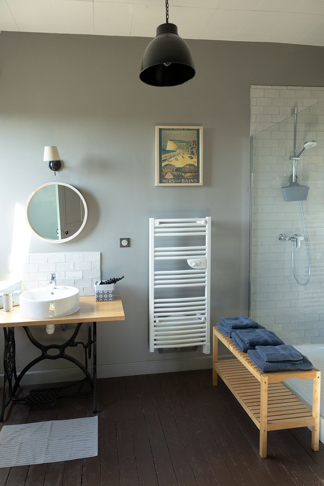 A bathroom with a round mirror, a sink, a towel rack, a shower, and a wooden bench.