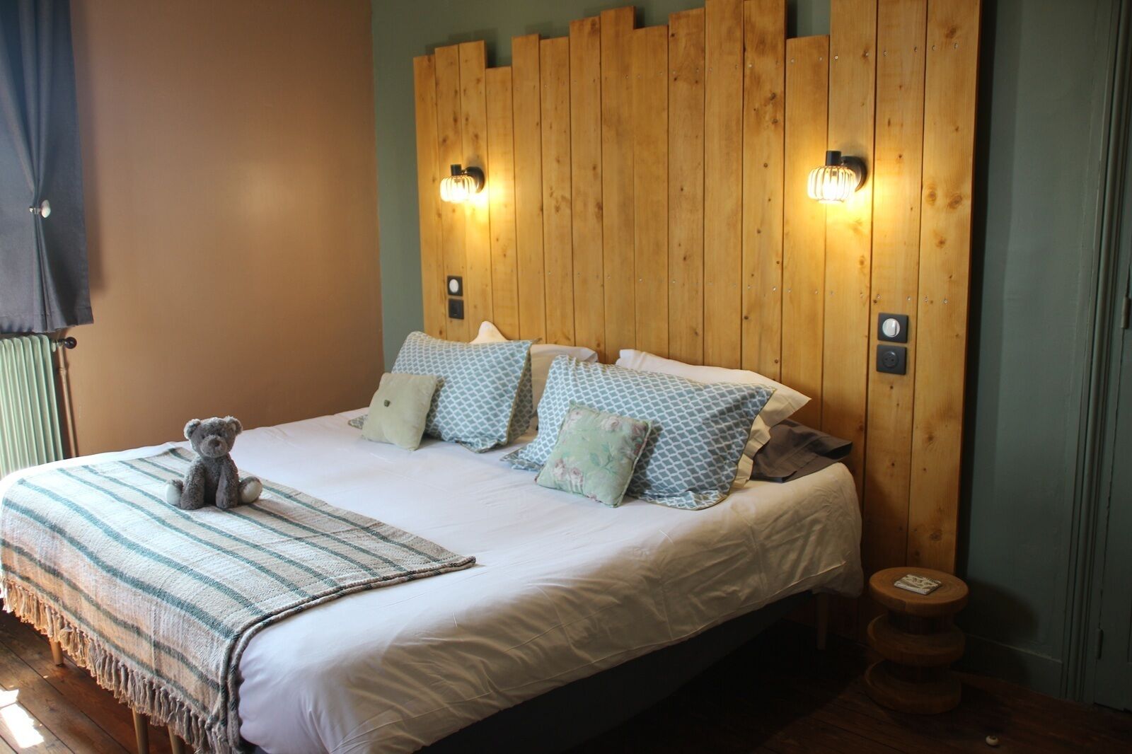A bed with a striped blanket, multiple pillows, a teddy bear, and wooden headboard with wall-mounted lights.