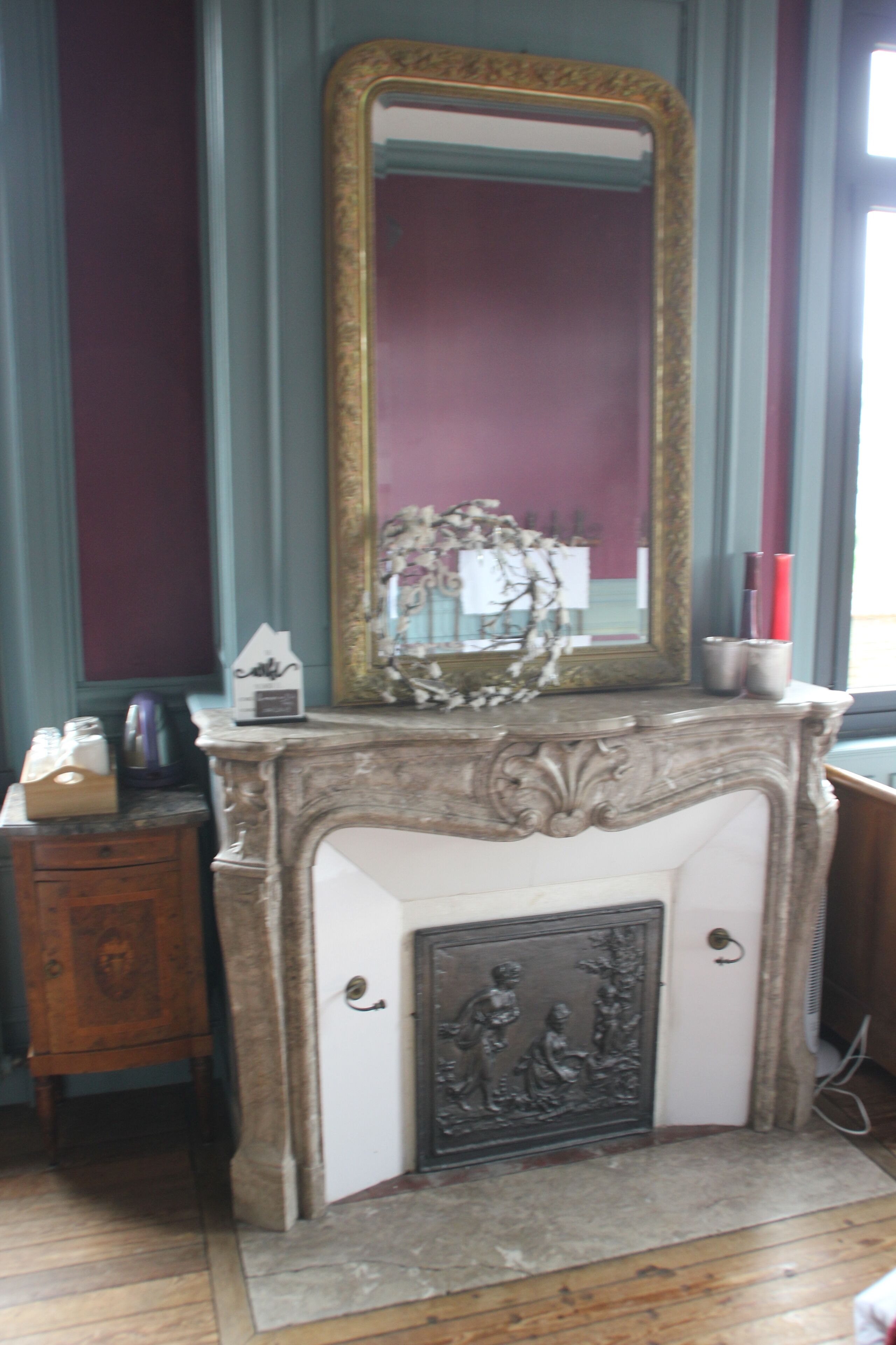 A room with a fireplace, a mirror, a small cabinet, and a window with curtains.