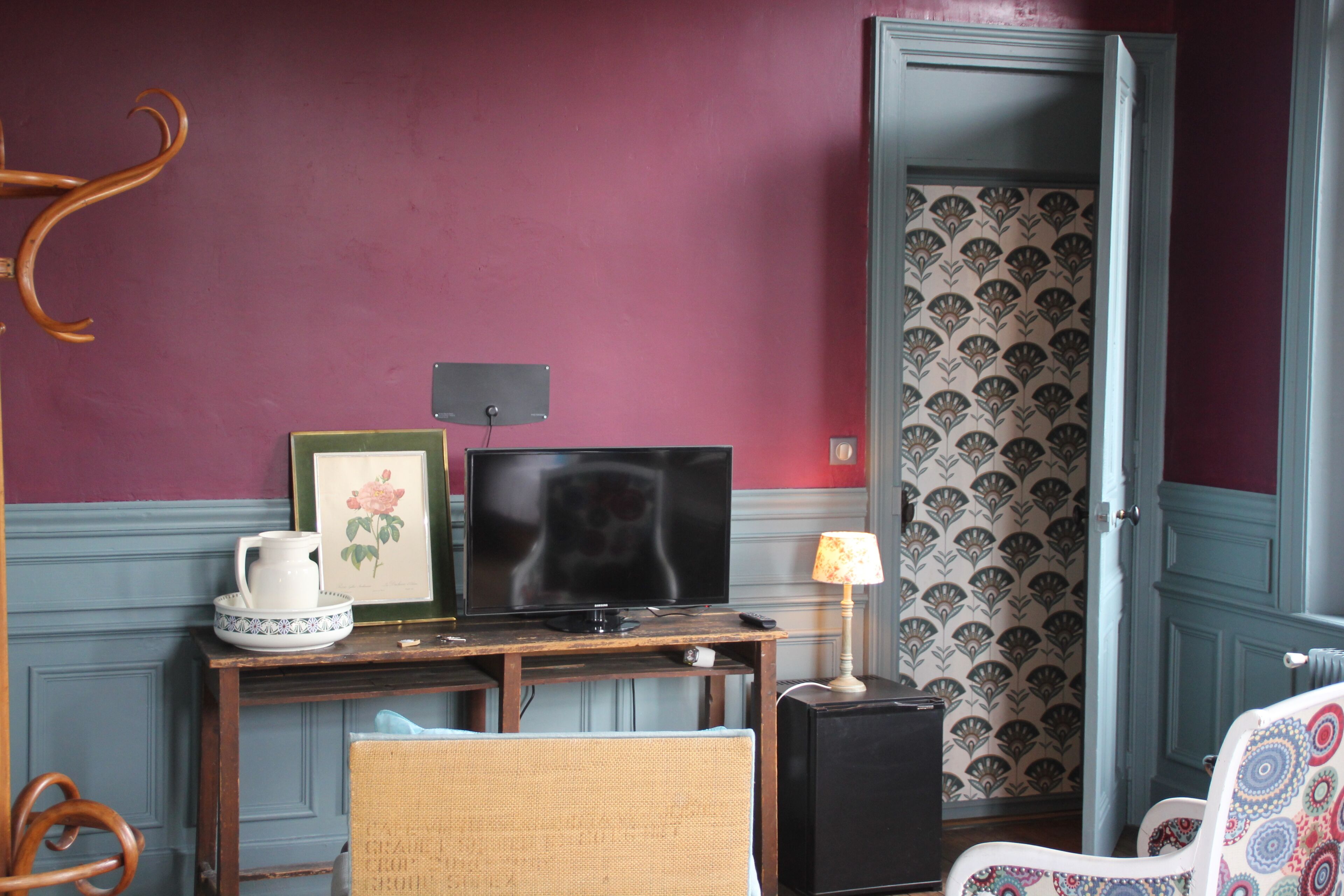 A room with a television on a wooden desk, a wicker chair, a patterned wallpaper, and a door with a decorative panel.