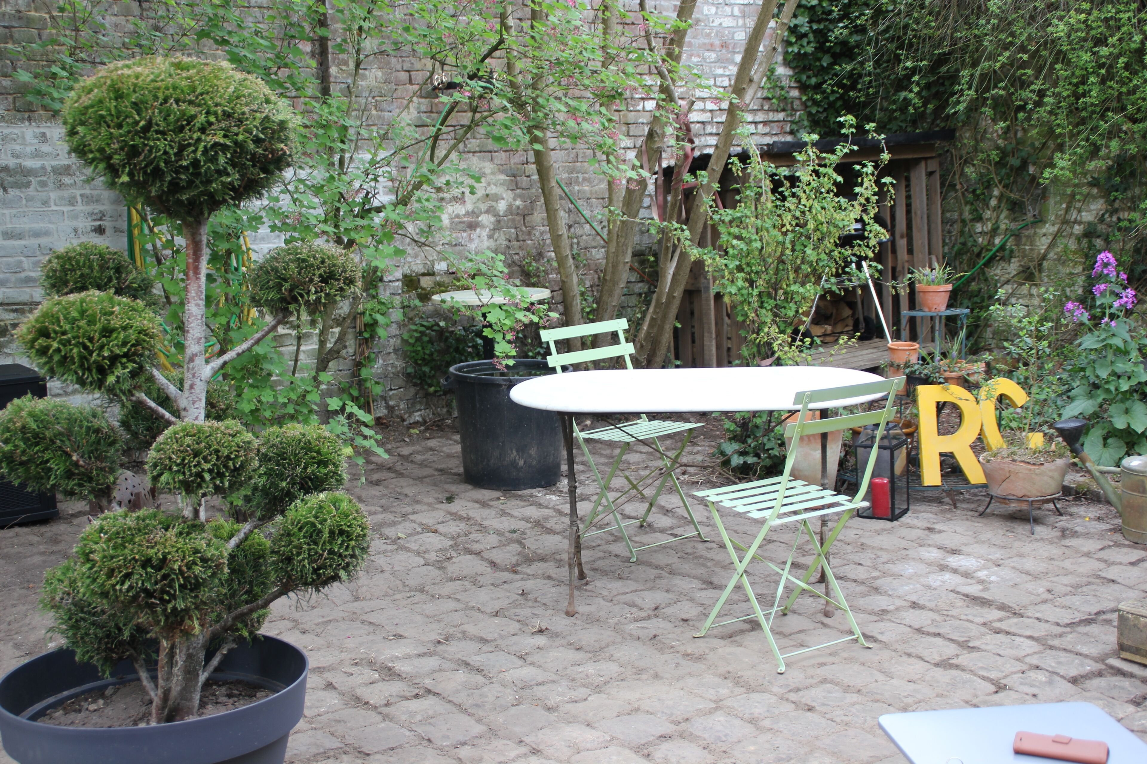 A patio with a round table and two green chairs, surrounded by potted plants and a brick wall.