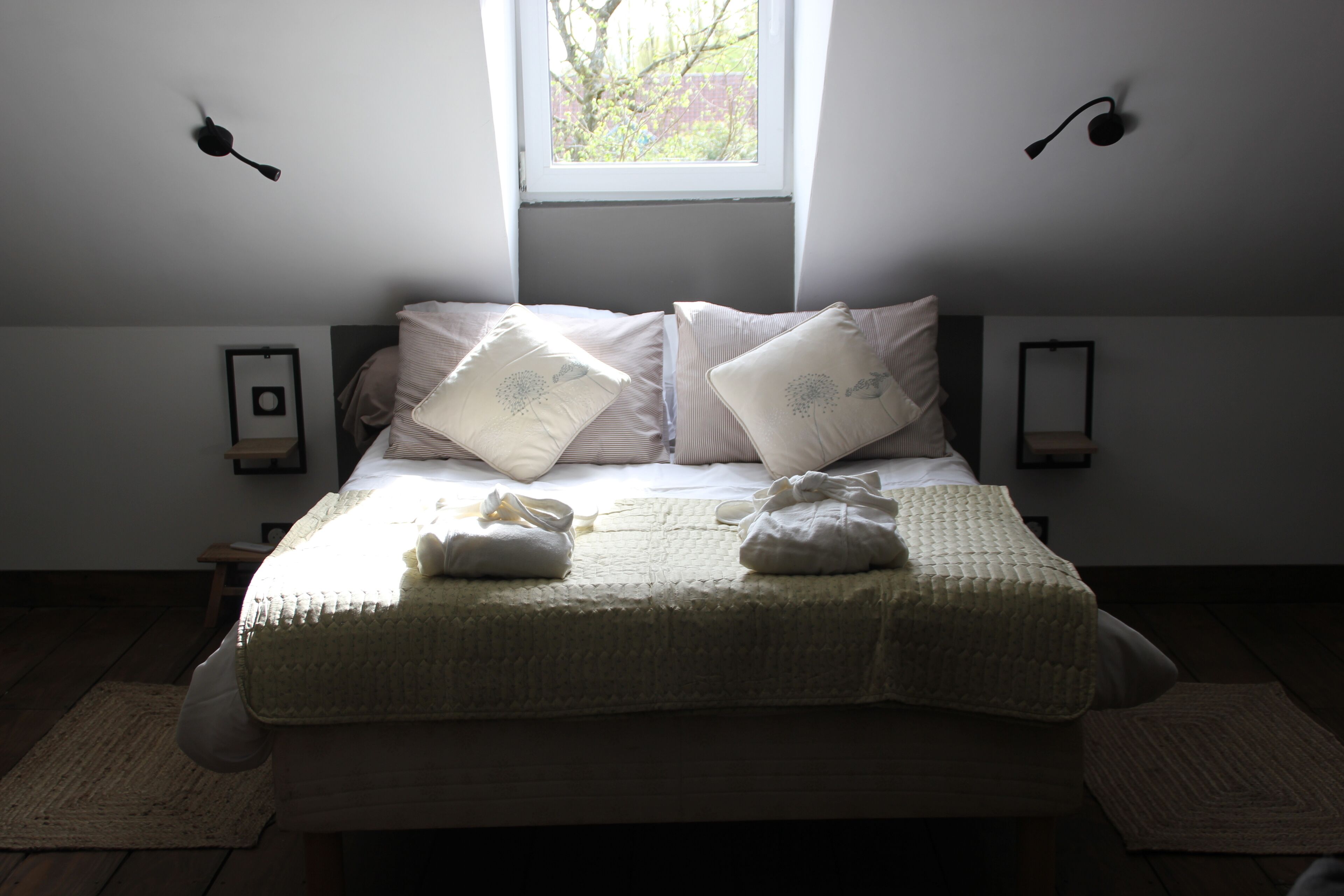 A neatly made bed with pillows and a blanket, a window with a view, and wall-mounted lights.