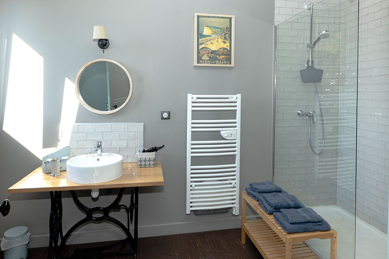A bathroom with a round mirror, white sink, towel rack, shower, and a bench.