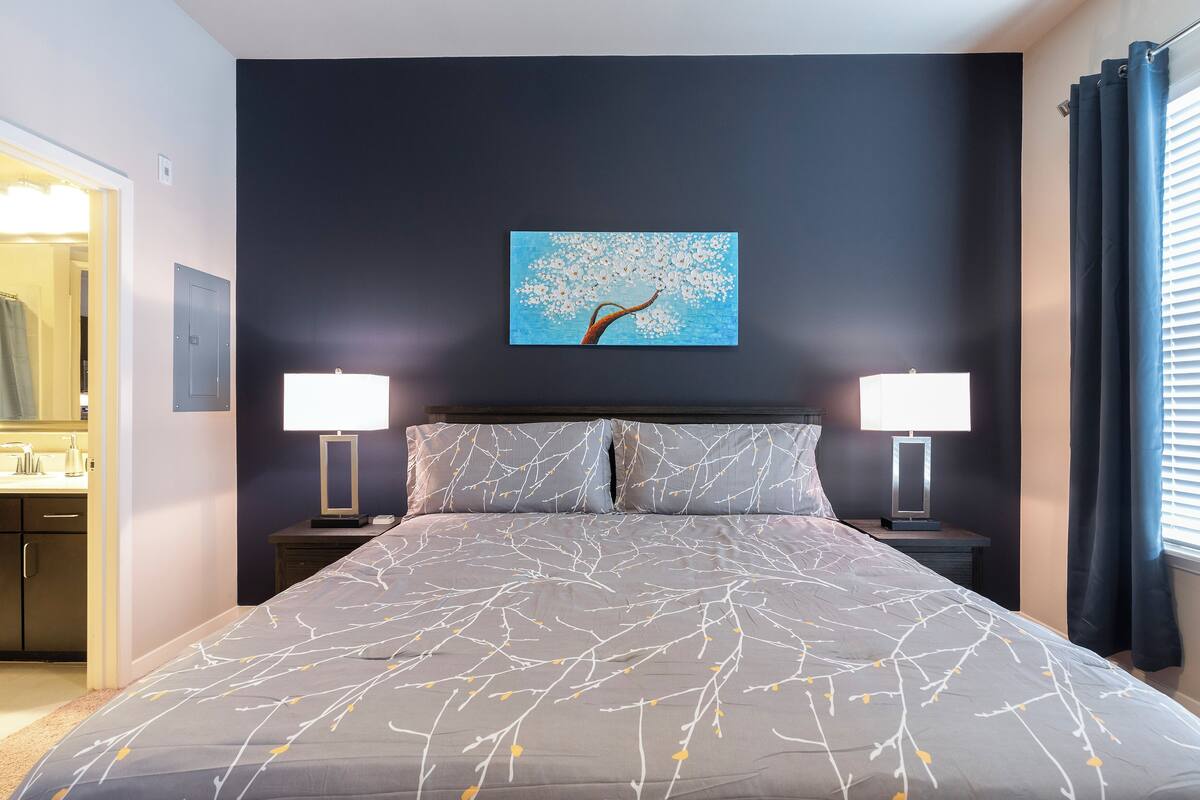 A bedroom with a bed, two bedside tables with lamps, a painting on the wall, and a window with curtains.