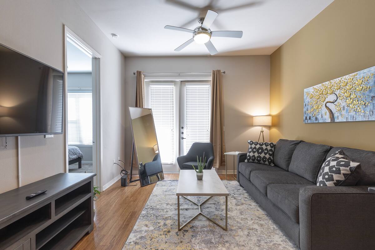 A modern living room with a flat-screen TV, a ceiling fan, a gray sofa, a coffee table, and a painting on the wall.