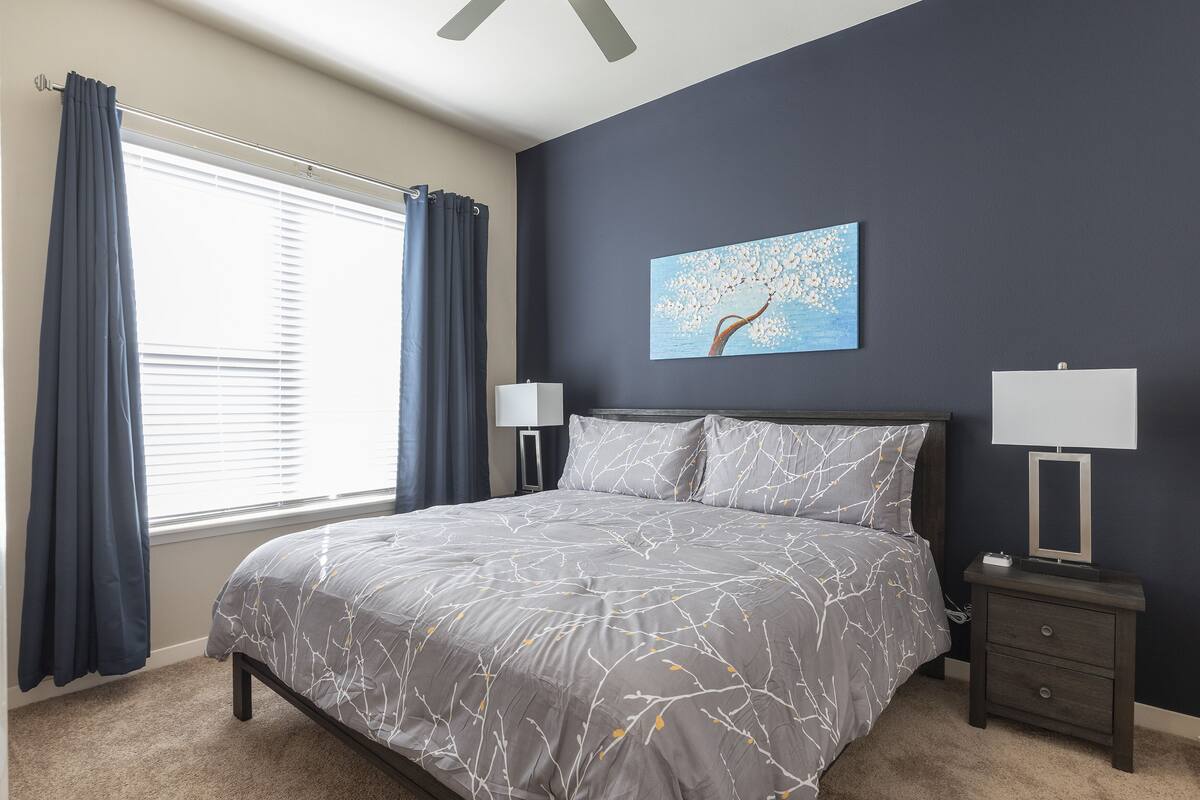 A bedroom with a bed, two bedside tables, a nightstand, a window with curtains, and a wall-mounted painting.