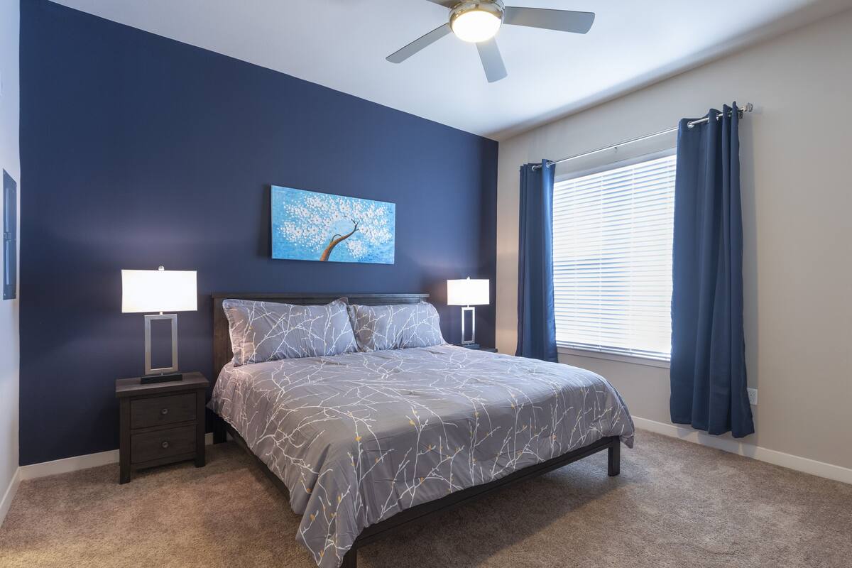 A bedroom with a bed, nightstands, a ceiling fan, a window with curtains, and a wall painting.