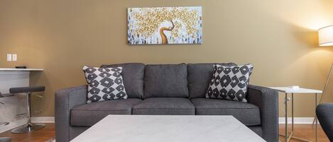 Una sala de estar moderna con un sofá gris, cojines estampados, una mesa de centro blanca y un cuadro en la pared.